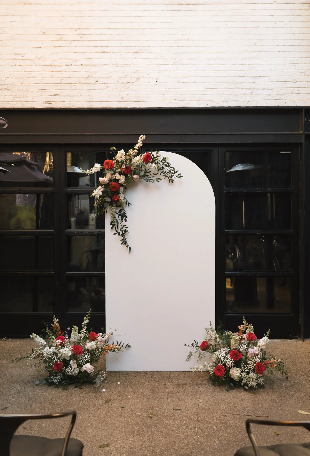White wedding arch with crimson roses and greenery against black doors in the Maketto courtyard